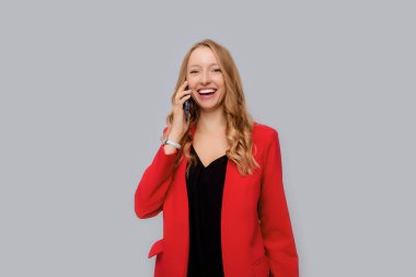 Gülümseyen mutlu iş kadını cep telefonuyla konuşuyor, bir telefon üzerine gülüyor, gri arka planda kırmızı ceketle dikiliyor. İş ve teknoloji.