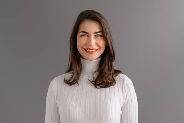 Beyaz kazaklı güzel bir esmer kadın stüdyoda gri bir arka planda durup gülüyor. Güzellik ve zarafet