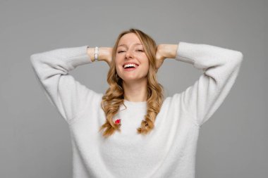 Yüzünde mutlu bir gülümsemeyle mutlu ve güzel bir genç bayan. Tasasız genç kadın ellerini başının üstüne koyup gri arka planda tasasız bir poz veriyor.