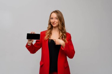 İş ve teknoloji. Güzel ve kendine güvenen bir iş kadını, gri arka planda kırmızı ceketli, yatay, boş akıllı telefon ekranını işaret ediyor.