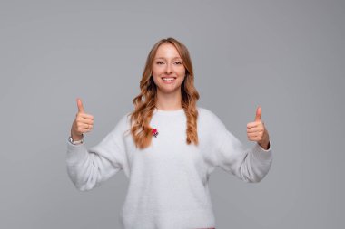 İyi iş çıkardın. Gülümseyen mutlu sarışın kadın başparmağını kaldırıyor ve mutlu görünüyor. Kızlar bunu onaylar, iltifat eder, beyaz süveter giyer, gri arka plan