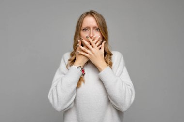 Şok olmuş genç sarışın bir kadının portresi. Ağzı kaplayan ve kameraya korkmuş bir ifadeyle bakan, korkunç bir şey olmasını bekleyen, beyaz süveter giyen, gri arka planda duran.