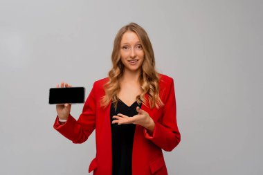 Güzel ve kendine güvenen iş kadını yatay olarak boş akıllı telefon ekranı gösteriyor, mobil uygulama gösteriyor, gri arka planda kırmızı ceketle duruyor.