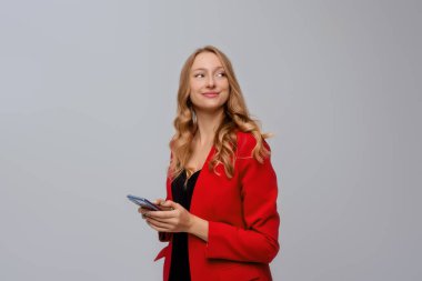 Başarılı genç girişimci kadın portresi, cep telefonunu tutuyor, gülümsüyor ve mutlu bir yüz ifadesi takınıyor, gri arka planda duruyor.