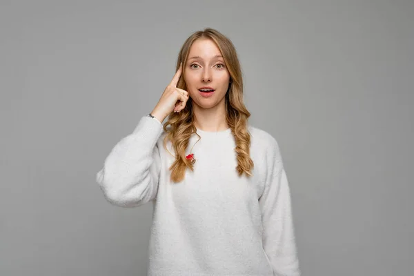 Portrait of surprised smiling young woman dressed cozy white sweater, twisting her index finger at temple, asking to think carefully or saying: What a crazy idea. Are you an idiot. Are you kidding me