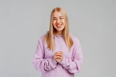 Gülümseyen güzel sarışın kızın portresi, kameraya mutlu bakıyor, arkadaşça gülümsüyor ve gri arka planda modaya uygun kapüşonlu bir şekilde dikilmiş yardım etmeye hazır.
