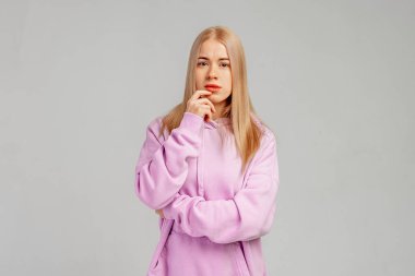 Düşünceli yüzlü genç sarışın kadın çeneye dokunuyor, düşünüyor, karar veriyor, gri arka planda modaya uygun mor kapüşonluyla dikiliyor.