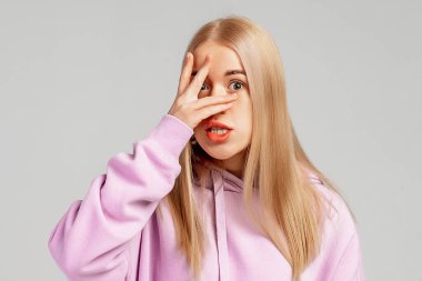 Korkmuş sarışın kızın yüzünü örten, endişeli ve şok olmuş, bir şeyden korkan, sıradan kıyafetler giyen, gri arka planda duran