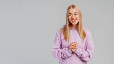 Gülümseyen güzel sarışın kızın portresi, kameraya mutlu bakıyor, arkadaşça gülümsüyor ve gri arka plan üzerinde modaya uygun bir kapüşonluyla duruyor yardıma hazır, boşluğu kopyalıyor.