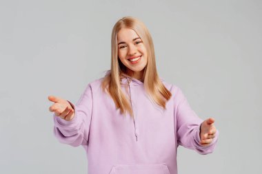 Moda kapüşonlu çekici kadın modelin portresi, kollarını kameraya uzatarak sarılmak, kucaklamak, kucaklamak, bir şey almak için tepki vermek, gri arka planda durmak.
