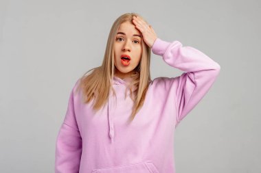 Eyvah, eyvah. Genç sarışın kadın menekşe kapüşonlu giyer, gri arka plana karşı durur, hata için elleri kafasında şaşırır, hatayı hatırla. Unuttum, kötü hafıza konsepti