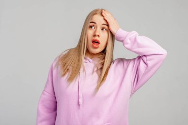 Eyvah, eyvah. Genç sarışın kadın menekşe kapüşonlu giyer, gri arka plana karşı durur, hata için elleri kafasında şaşırır, hatayı hatırla. Unuttum, kötü hafıza konsepti