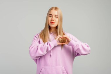 Çekici, neşeli bir kızın portresi, kalp şeklinde amour gösteriyor, gri arka planda modaya uygun bir kapüşonluyla duruyor. Aşkın işareti, ahenk, minnettarlık, hayırseverlik. Duygular ve duygular kavramı