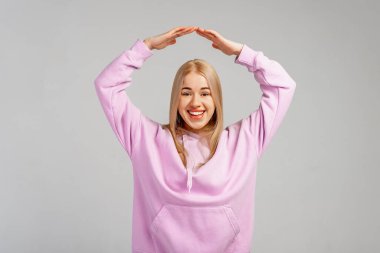 Gülümseyen, genç, sarışın bir kadın elleriyle çatı jesti, güvenlik sembolü, koruma, yeni ev, satın alma hayali. Evini koru.