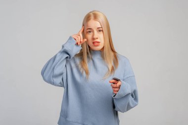 Hoşnutsuz genç, sarışın bir kadının görüntüsü kızgın ya da yargılayıcı bir şekilde kameraya işaret parmaklarıyla bakıyor, mavi eşofman altı giyiyor, gri arka planda duruyor. Vücut dili kavramı