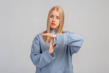 Kafası karışmış sarışın kadın duraklatıyor ya da mola işareti yapıyor, beklemek istiyor, son teslim tarihine üzülüyor, gri arka planda duruyor. Vücut dili kavramı. Zaman sınırı