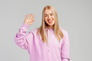 Gülümseyen mutlu kızın portresi arkadaşça el sallıyor, merhaba diyor, selam veriyor, selam veriyor, mor kapüşonlunun içinde gri arka planda duruyor.