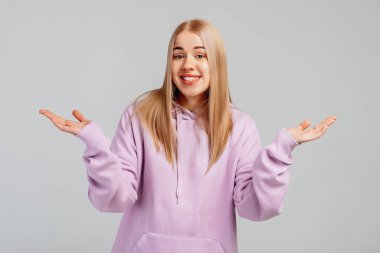 Genç sarışın bir kadın yanılıyor, ellerini açıyor, ah, ah, ah, ah, ah, ah, jest, kafası karışmış görünüyor, gri arka planda duruyor.