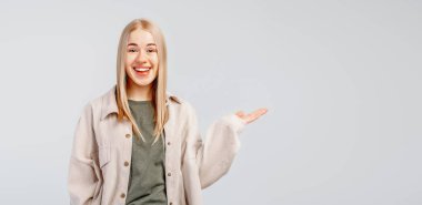 Modaya uygun tişörtlü, güler yüzlü, şık sarışın kız elinde gri arka planda duran bir şey gösteriyor. Boşluğu sağ tarafa kopyala