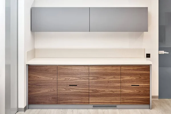 Çağdaş mutfak mobilyaları, minimalist tarzda ceviz ağacı ahşap ve gri renkte. Ofiste akrilik katı yüzey tezgahı var.