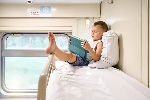 ブロンド小さな男の子はタブレットPcを介して漫画を見て 午前中に電車のコンパートメントで上の棚の上に柔らかい枕にもたれて — ストック写真