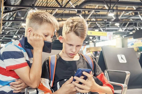 集中ブロンドの兄弟はゲームをプレイし スマートフォンを使用して興味を共有し 空港のアームチェアで待っているホールに座っています — ストック写真