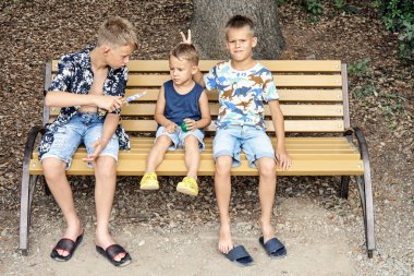 Ağabeyim, tahta bankta oturan küçük çocukla alay ediyor. Anaokulu öğrencisi yaz tatilinde parkta uzun bir yürüyüşten sonra kardeş şakalarından dolayı üzgün.