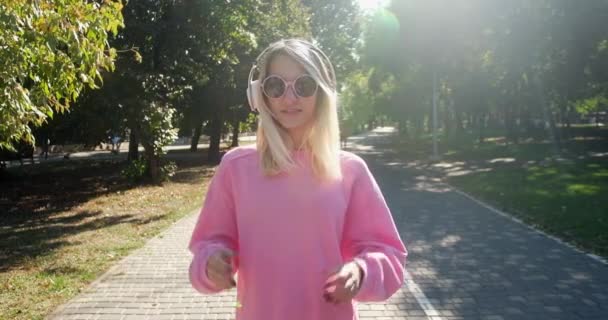 Junge Frau posiert mit Kopfhörern und Sonnenbrille im Park — Stockvideo