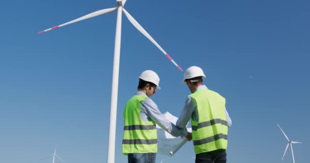 Engenheiros olham para o projeto desenhando contra moinhos de vento rotativos — Vídeo de Stock