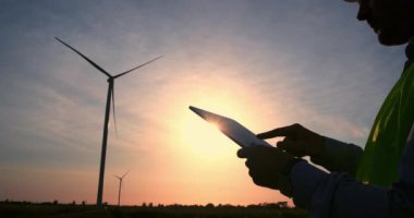 Bir mühendis rüzgar jeneratörünün işleyişini bir bilgisayar aracılığıyla ayarlar. Gün batımında bir rüzgar jeneratörünün arka planında elinde tabletle gezen özel elbiseli bir adamın silüeti.. 