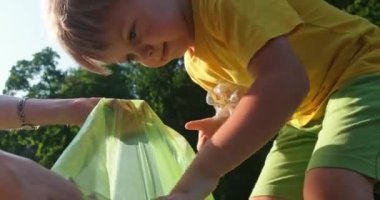 Sarışın küçük çocuk annesinin parkta plastik şişe toplamasına yardım ediyor.