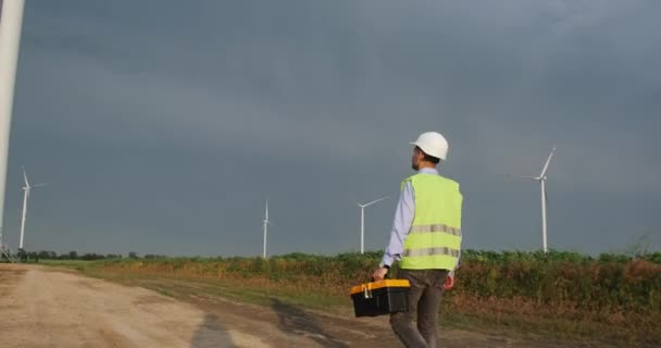 Electrician goes along path past high windmills in field — Vídeo de Stock