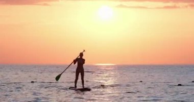 Genç kadın ayakta kürek çekiyor, günbatımının keyfini çıkarıyor.