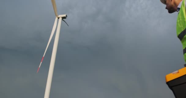 Man electrician looks at high windmill under gloomy sky — Vídeo de Stock