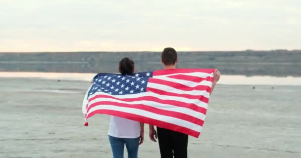 Man och kvinna håller och lyfter amerikansk flagga på flodstranden — Stockvideo