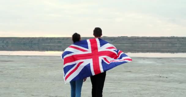 Pareja joven se une a las manos levantando bandera británica en la orilla del río — Vídeos de Stock
