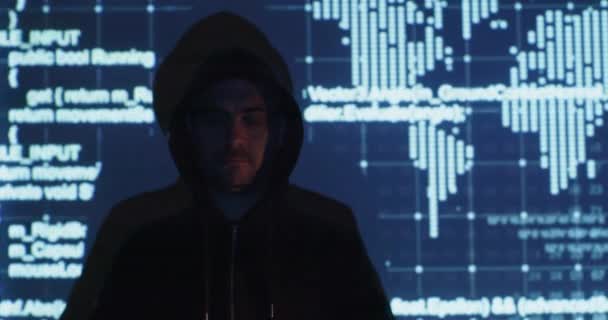 Hacker in black hoodie stands against digital screen — Stock Video