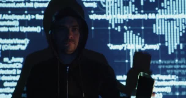 Hacker in hoodie shows smartphone and smiles near screen — Stockvideo