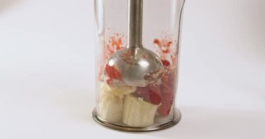 Housewife whips fresh banana and strawberry with blender in plastic container on white background. Preparation of smoothie at home extreme closeup
