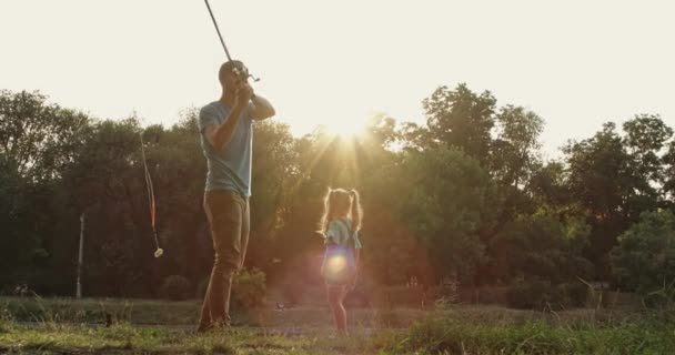お父さんは魚を捕まえるために釣竿を川に投げ 小さな娘は公園で助けます お父さんとともにブロンド女の子過ごす時間で日没遅い動き — ストック動画