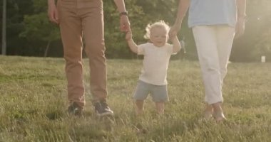 Küçük çocuk halka açık bir parkta ailesinin elini tutarak yürümeyi öğreniyor. Aile, sıcak yaz gününde oğluyla birlikte yeşil park bölgesinde yürüyor.