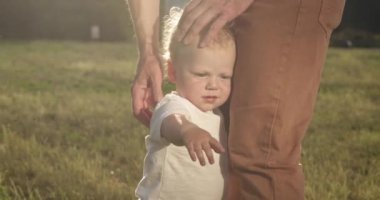 Cute toddler boy hugs father leg in park and curiously looks around. Parent carefully supports little son touching head and back closeup
