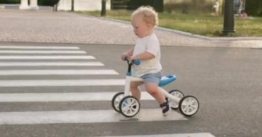 Cute toddler boy rides bicycle on empty roadway and crosses pedestrian crossing. Blond toddler learns to ride bike outdoors on city street