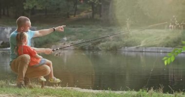 Dad and little daughter sit on park river bank fishing at back sunset. Father shows fish in water to daughter against walking people slow motion