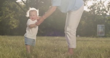 Caring mother teaches little toddler boy to walk barefoot in park. Toddler sits on ground after making few steps with mother support