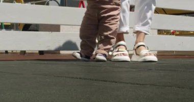 Mother teaches toddler son walking outdoors on sunny day. Mom and child slowly walk together on asphalt path. Little kid learns to walk