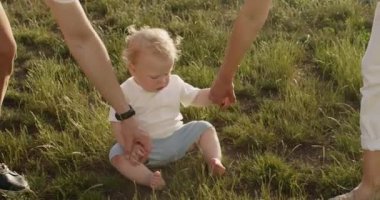 Parents take hands of little son and carefully put baby boy on feet. Loving parents teach little toddler boy to walk on park lawn closeup