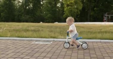 Blond little boy rides fast children bike in public park on pavement road. Active toddler rides small bicycle in green park against trees