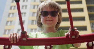 Güneş gözlüklü gülümseyen çocuk çocuk çocuk parkında eğleniyor. Sarışın anaokulu öğrencisi kırmızı tırmanma halatına tırmanıyor ve kameraya heyecanlı bir şekilde bakıyor.