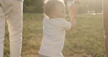 Curly toddler boy walks on park field with caring parents on sunny day. Family enjoys free time in park. Parents teach son to walk on meadow back view